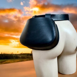 Puede incluir: Una riñonera de cuero negro con un cinturón negro ancho. La riñonera está unida a un torso de maniquí blanco.