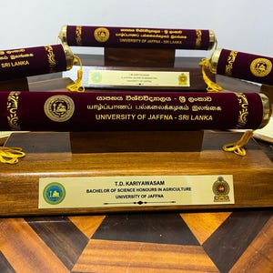 May include: A display of maroon diploma tubes with gold accents, each bearing the University of Jaffna name in English and another language. The diplomas are mounted on a wooden base with nameplates, including one for T.D. Kariyawasam.