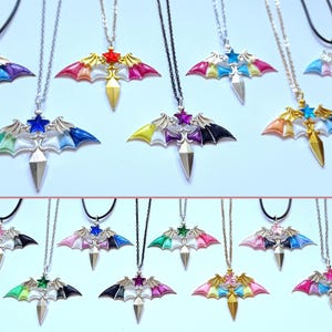 Op de afbeelding: Een verzameling kleurrijke hangerkettingen. Elke ketting heeft een vleermuisvleugelontwerp met een ster en een diamantvormige hanger. De vleugels zijn geschilderd in verschillende kleuren, waaronder roze, blauw, geel en groen. De kettingen hebben zwarte of zilveren kettingen.