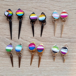 Op de afbeelding: Een verzameling handgemaakte oorbellen met verschillende LGBTQ+ pride vlag ontwerpen. Elke oorbel heeft een rond, kleurrijk ontwerp met een spike hanger. De oorbellen zijn in verschillende kleuren, waaronder zwart, goud en zilver.