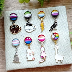 Puede incluir: Un conjunto de 10 pines de esmalte con varios diseños, que incluyen un gato negro en una taza de café, un cuervo, un fantasma, un conejito, una llama y diferentes banderas del orgullo. Los pines están unidos a una tabla blanca.