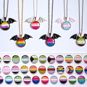 Puede incluir: Una colección de collares y pines redondos con varios diseños de banderas del orgullo. Los collares tienen un colgante de ala de murciélago. Los pines y colgantes muestran una variedad de colores, incluyendo arcoíris, rosa, azul, amarillo y negro, sobre un fondo blanco.