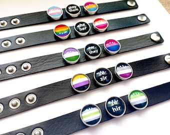 May include: Six black leather bracelets with silver studs and colorful round buttons. Each button features a different pride flag and text, including "she her", "they them", "ze zir", and "ze hir".