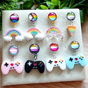 May include: A collection of colorful earrings featuring rainbow and video game controller designs. The earrings have various pride flag color schemes. The earrings are displayed on a light-colored, textured surface.