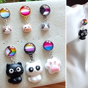 Könnte beinhalten: Ein Set mit sechs Paar Ohrringen mit verschiedenen Pride-Flaggen und niedlichen Tieranhängern. Die Ohrringe sind aus Glascabochons, Metallanhängern und Metallsteckern gefertigt. Die Flaggen umfassen die lesbische, bisexuelle, transgender, pansexuelle, asexuelle und genderfluide Flagge. Die Tieranhängern umfassen Katzen, einen Pfotenabdruck und eine schwarze Katze.