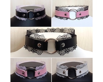 May include: A selection of chokers in black, pink, and white. Each choker has a leather band, a large silver ring, and delicate lace trim. The chokers are displayed on a mannequin head.