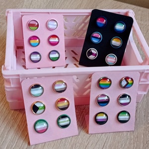 May include: Six sets of pride flag earrings displayed on pink card stock. Each set features two earrings with a different pride flag design. The flags include the lesbian, bisexual, transgender, non-binary, pansexual, and genderfluid flags.