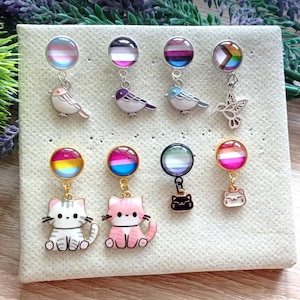 Op de afbeelding: Een collectie van acht hangende oorbellen met ronde cabochons in diverse pride-vlagpatronen. De bovenste rij bevat vier vogelbedels, terwijl de onderste rij vier kattenbedels toont in verschillende kleuren, waaronder wit, roze en zwart.