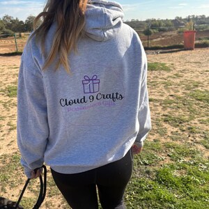 May include: A grey hoodie with a drawstring hood and the text "Cloud 9 Crafts Personalized Gifts" in purple. The hoodie is worn by a person walking a dog on a sunny day.