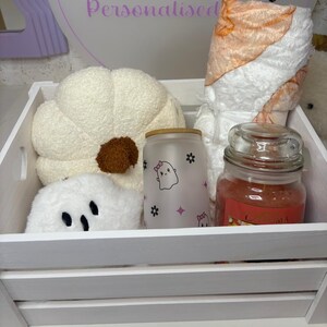 May include: A white wooden crate containing a cream-coloured plush pumpkin, a white plush ghost, a frosted glass tumbler with a ghost and flower print, and a rolled orange and white patterned blanket next to a jar candle with a red label.