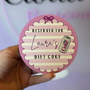 May include: A circular coaster with a scalloped edge featuring a pink and white horizontal striped pattern. The text reads RESERVED FOR Laura's DIET COKE, accompanied by a small illustration of a silver Diet Coke can and a purple bow icon.