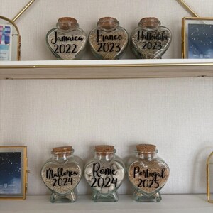 May include: Six heart-shaped glass jars with cork stoppers, filled with sand or small stones. Each jar is labeled with a destination and year: Jamaica 2022, France 2023, Halkidiki 2023, Mallorca 2024, Rome 2024, and Portugal 2025. The jars are displayed on a white shelf.