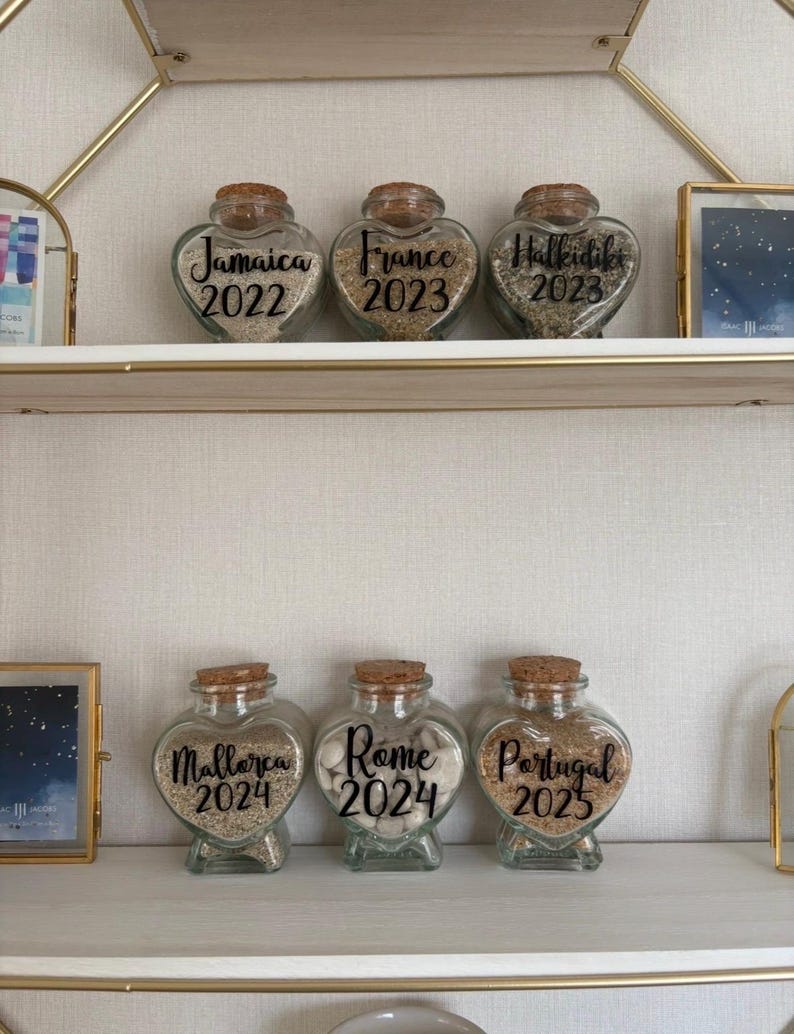 May include: Six heart-shaped glass jars with cork stoppers, filled with sand or small stones. Each jar is labeled with a destination and year: Jamaica 2022, France 2023, Halkidiki 2023, Mallorca 2024, Rome 2024, and Portugal 2025. The jars are displayed on a white shelf.