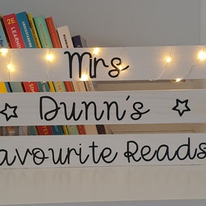 May include: White wooden crate with a sign that says "Mrs. Dunn's Favourite Reads" and a string of fairy lights. The crate is filled with books.