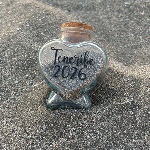 May include: A clear, heart-shaped glass bottle filled with small, dark grains. The bottle has a cork stopper and the words "Tenerife 2026" are written on the front in a script font. The bottle sits on a bed of dark sand.