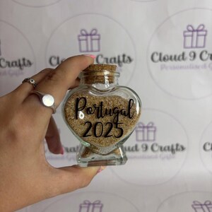 May include: A heart-shaped glass jar filled with sand, labeled "Portugal 2025". The jar has a cork stopper and is held in a hand. The background features a repeating pattern of gift box graphics.