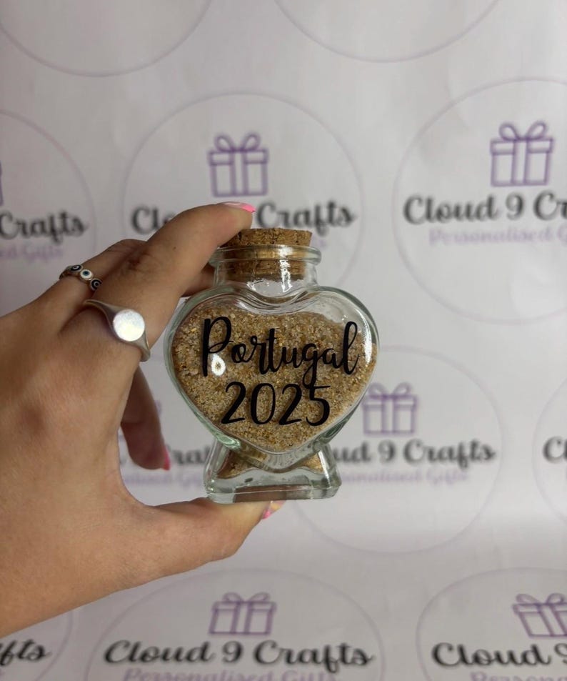 May include: A heart-shaped glass jar filled with sand, labeled "Portugal 2025". The jar has a cork stopper and is held in a hand. The background features a repeating pattern of gift box graphics.