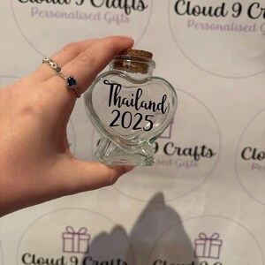 May include: A clear, heart-shaped glass jar with a cork stopper. The jar is labeled "Thailand 2025" in black script. The background features the "Cloud 9 Crafts" logo and a hand holding the jar.