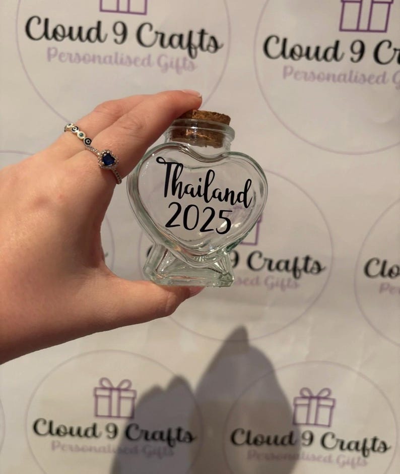 May include: A clear, heart-shaped glass jar with a cork stopper. The jar is labeled "Thailand 2025" in black script. The background features the "Cloud 9 Crafts" logo and a hand holding the jar.