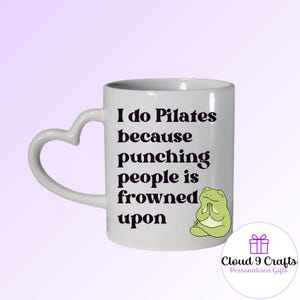 May include: White ceramic mug with a heart-shaped handle. The mug features the text "I do Pilates because punching people is frowned upon" in black, along with a cartoon frog illustration. A logo is visible at the bottom.