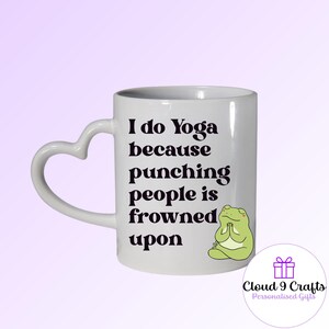 May include: White ceramic mug with a heart-shaped handle. The mug features the text "I do Yoga because punching people is frowned upon" and a cartoon frog. A purple logo is on the bottom right.