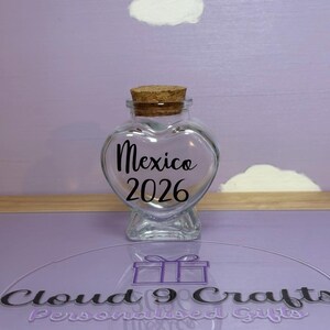 May include: A clear, heart-shaped glass jar with a cork stopper. The jar has the word "Mexico" and the year "2026" in black script. The background is a light purple with white cloud-shaped decorations.