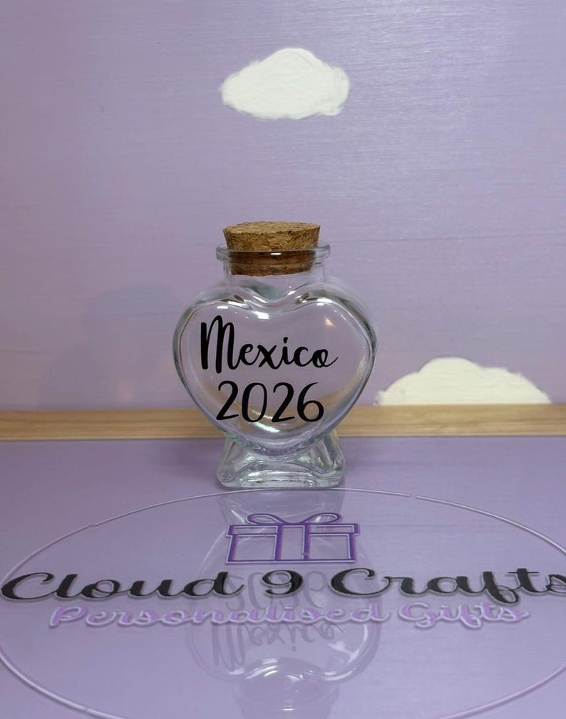 May include: A clear, heart-shaped glass jar with a cork stopper. The jar has the word "Mexico" and the year "2026" in black script. The background is a light purple with white cloud-shaped decorations.