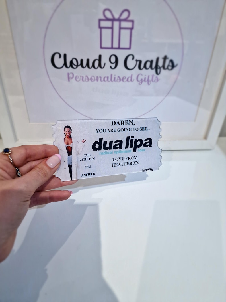 Pu&ograve; includere: Un biglietto bianco con una foto di Dua Lipa, il testo "DAREN, YOU ARE GOING TO SEE... dua lipa", "TUE 24TH JUN SPM ANFIELD", "radical optimism", e "LOVE FROM HEATHER XX".