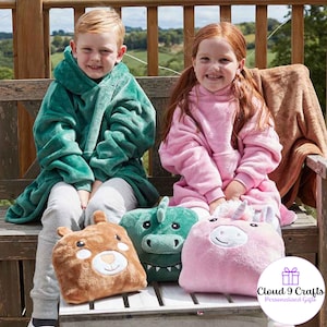 May include: Two children wearing hooded blankets, one green and one pink, sit on a wooden bench. They hold plush animal pillows: a brown bear, a green dinosaur, and a pink unicorn. The image includes the text "Cloud 9 Crafts".