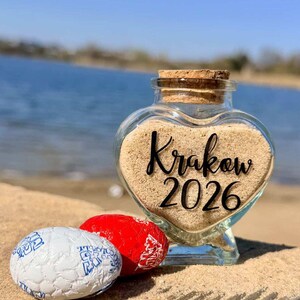 May include: A heart-shaped glass jar filled with sand, featuring the text "Krakow 2026". Two wrapped candies, one red and one white with blue designs, sit in front of the jar. The scene is set on a sandy surface near water.