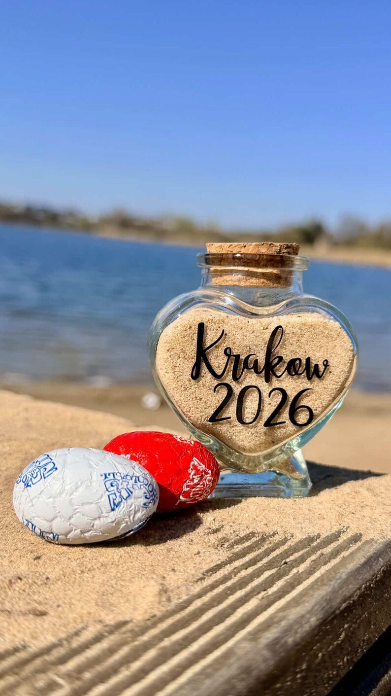 May include: A heart-shaped glass jar filled with sand, featuring the text "Krakow 2026". Two wrapped candies, one red and one white with blue designs, sit in front of the jar. The scene is set on a sandy surface near water.
