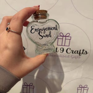 May include: A clear glass heart-shaped bottle with a cork stopper. The bottle has the words "Engagement Sand" in black script. The bottle is held up against a white background with purple gift box graphics.