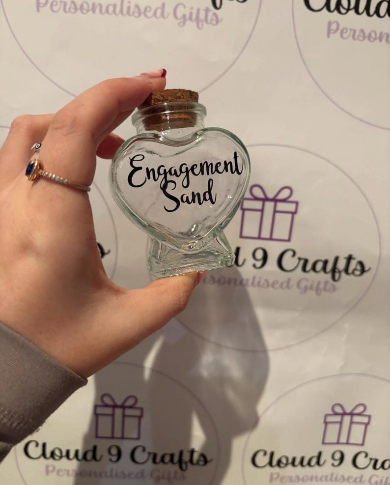 May include: A clear glass heart-shaped bottle with a cork stopper. The bottle has the words "Engagement Sand" in black script. The bottle is held up against a white background with purple gift box graphics.