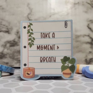 May include: A blue and white sticker with a paperclip, a potted plant, a candle, and the text "Take a moment & breath".