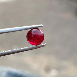 May include: A round, polished ruby gemstone held by silver tweezers. The ruby is a deep, vibrant red color with visible internal patterns. The gemstone is the focal point, showcasing its smooth, reflective surface.