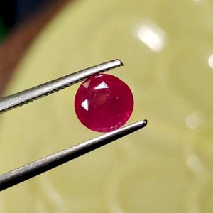 Puede incluir: Una piedra preciosa redonda, facetada y de color rojo intenso, sostenida por unas pinzas de metal plateado. La piedra muestra un tono carmesí brillante y saturado con reflejos de luz visibles en su superficie pulida, exhibiendo un corte circular claro.
