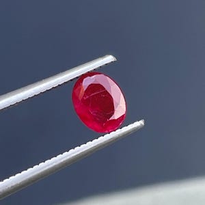 Może przedstawiać: Owalny, intensywnie czerwony kamień szlachetny trzymany przez pęsetę. Kamień jest gładki i wypolerowany, o głębokim czerwonym odcieniu.