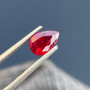 May include: A pear-shaped ruby gemstone with a deep red color. The gemstone is held in tweezers.