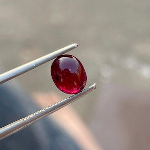 Pode incluir: Um rubi oval polido, segurado por uma pinça prateada. A gema é de cor vermelha profunda e tem uma superfície lisa e refletora. A pinça está em foco, enquanto o fundo está desfocado.