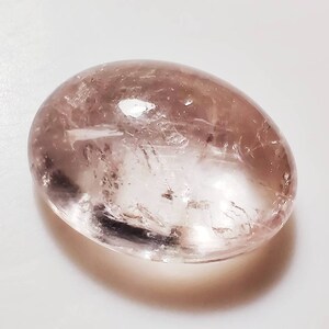 May include: A pale pink, oval-shaped gemstone with a smooth, polished surface. The stone has a translucent appearance with visible internal lines and inclusions.