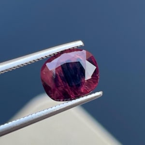 Può includere: Una gemma sfaccettata, di forma ovale e di colore rosso intenso, tenuta da una pinzetta d'argento. La gemma ha un interno scuro e una superficie lucida e riflettente, che mostra la sua chiarezza e taglio.