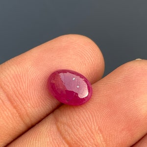 Puede incluir: Una gema de rubí de forma ovalada vibrante con un tono rojo intenso. La gema tiene una superficie lisa y pulida y está engastada en un corte cabujón.