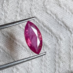 Op de afbeelding: Een edelsteen in marquise-slijpvorm met een levendige roze-rode tint, vastgehouden door een metalen pincet. De steen heeft meerdere facetten die het licht reflecteren, wat zorgt voor een schitterend uiterlijk met zichtbare interne insluitingen.