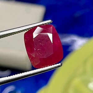 May include: A faceted, cushion-cut ruby gemstone held by silver tweezers. The ruby is a deep, saturated red colour with visible internal clarity. The background is a mix of blue and yellow.