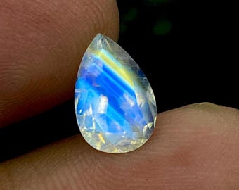 May include: A pear-shaped iridescent gemstone with a blue and yellow sheen. The gemstone is clear and has a faceted cut.