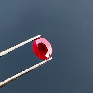 Puede incluir: Una gema de rubí facetada, de forma ovalada, sujeta con pinzas plateadas. El rubí es de un color rojo intenso y saturado, con reflejos internos visibles. Las pinzas se colocan sobre un fondo gris oscuro, resaltando el brillo de la gema.