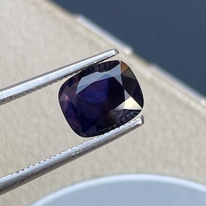 May include: A dark purple gemstone with a cushion cut. The gemstone is held in a pair of tweezers.