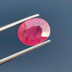 May include: A vibrant pink oval-shaped gemstone held by tweezers. The gemstone is faceted and has a smooth, polished surface.