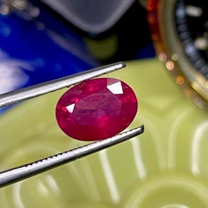 May include: A vibrant, oval-cut ruby gemstone with a deep red hue and faceted surface, held securely by a pair of silver-toned metal tweezers. The gemstone displays internal clarity and light reflection across its polished, symmetrical top surface.