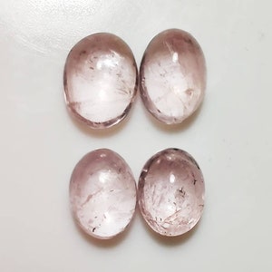 May include: Four oval-shaped, translucent gemstones with a soft pink hue. The stones are arranged in a square pattern against a white background. Each gem has internal inclusions, adding to their unique character.